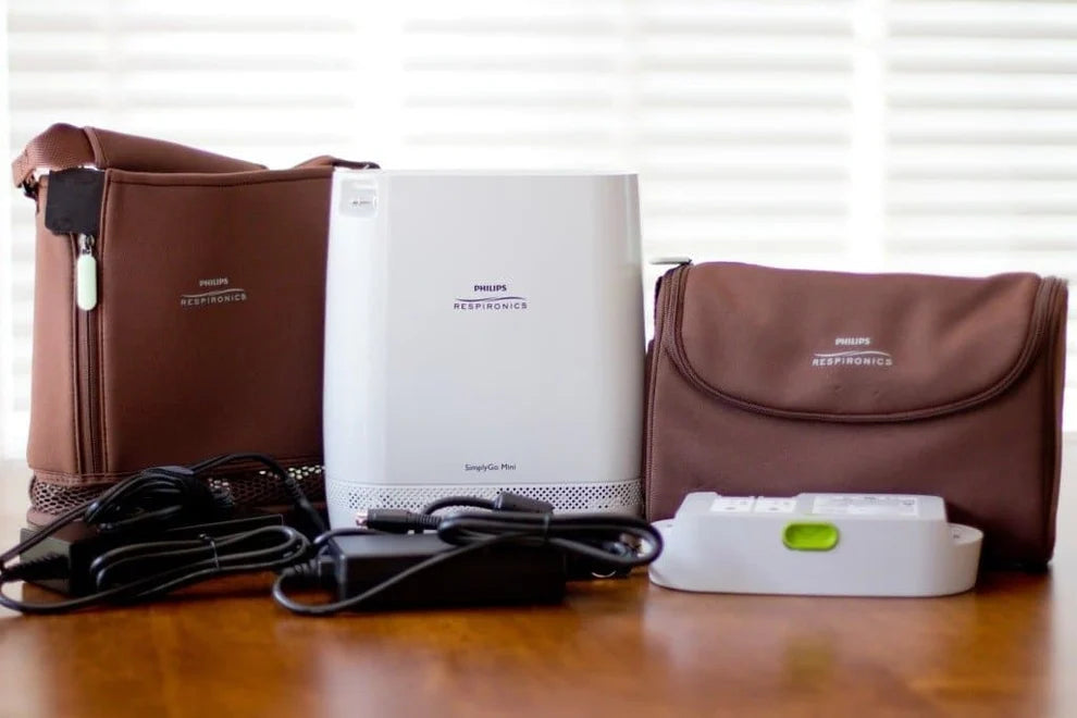 Philips Respironics Quiet Pro air purifier kit on a wooden table, including the white purifier, brown carrying cases, power adapter, and filter, with a window blind background.